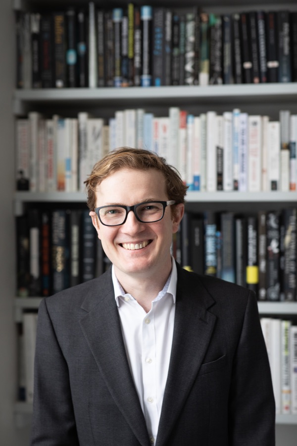 Henry smiling in a dark jacket in front of bookshelves